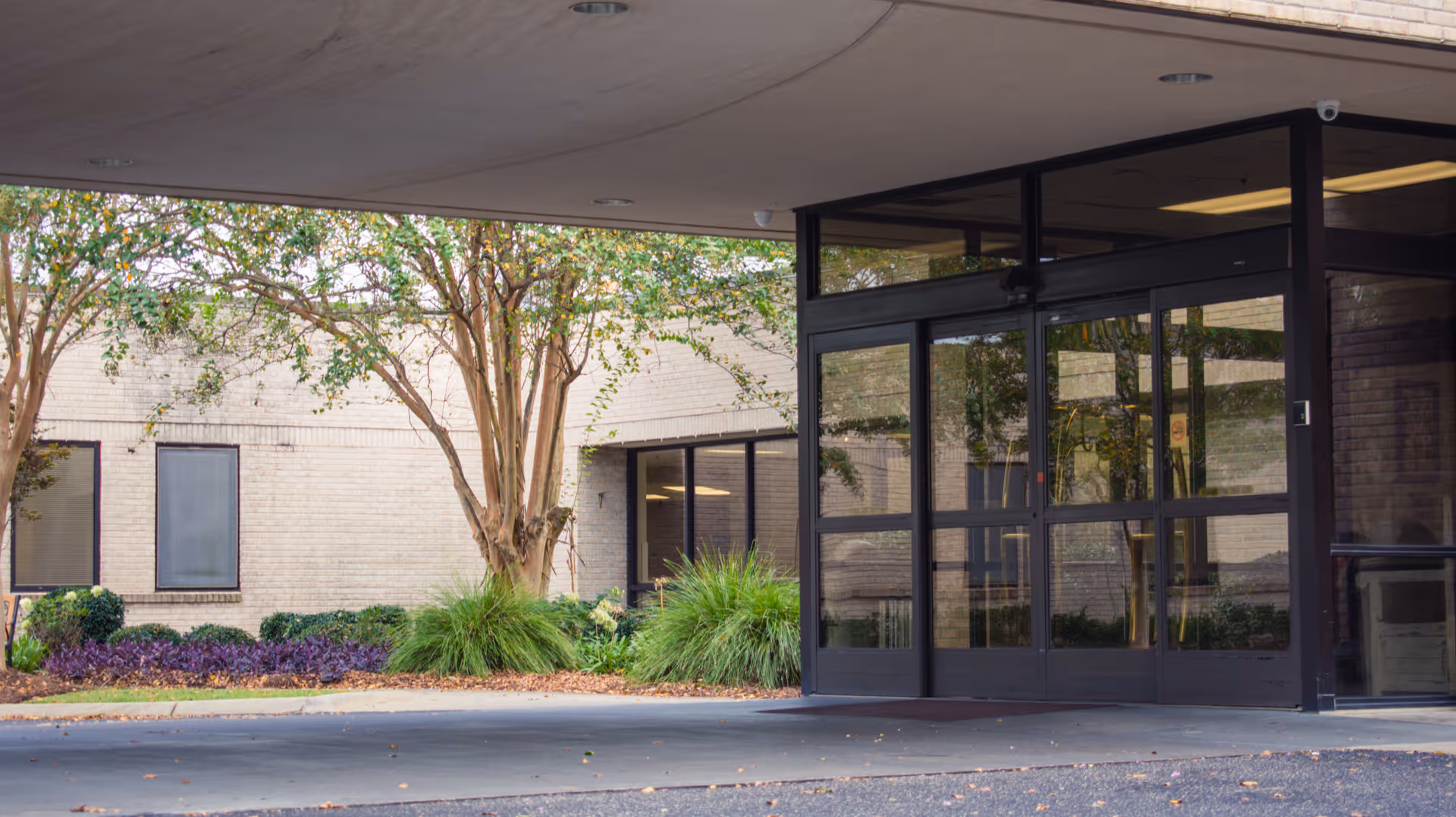 Entrance area of a healthcare facility with glass double doors, beige brick walls, and landscaping including trees and bushes under a covered drop-off area.