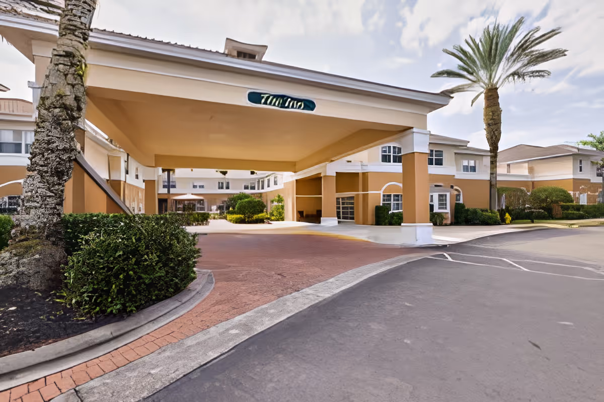 Entrance area of a senior living facility with a covered drop-off zone, palm trees, and a multi-story building in the background under a partly cloudy sky.