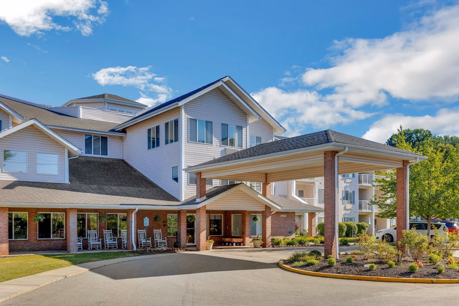 Exterior view of a multi-story senior living facility with a covered entrance, brick and white siding, rocking chairs on the porch, landscaped garden beds, and a clear blue sky with some clouds.