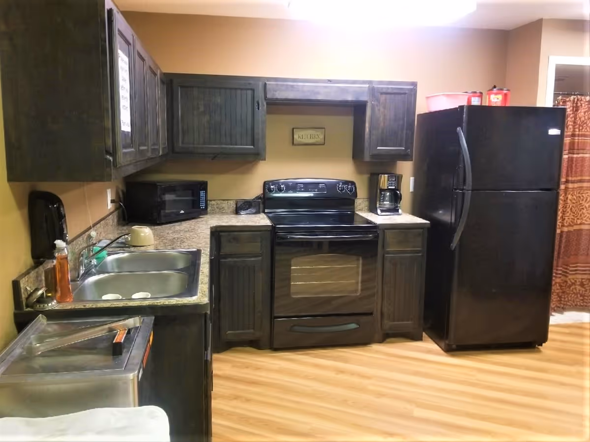 A small kitchen area with dark wooden cabinets, a black electric stove, a black refrigerator, a microwave, a coffee maker, a double sink, and a countertop with various items. The floor is light wood, and there is a beige wall with a small sign that says 'KITCHEN'.