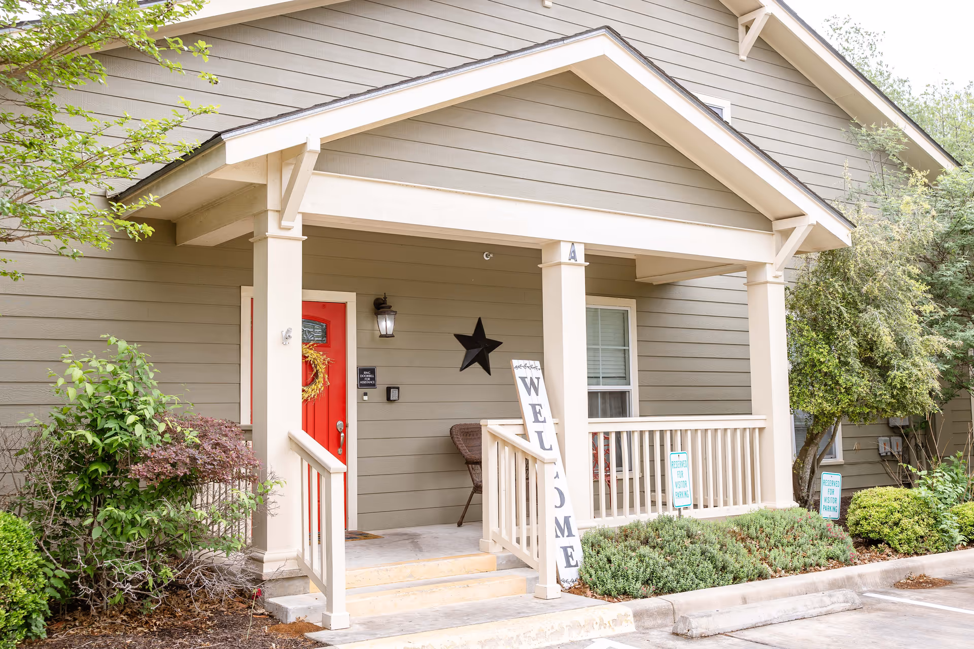 Exterior view of a building entrance with a covered porch, beige siding, a red door decorated with a wreath, a black star decoration on the wall, a chair on the porch, and a vertical wooden sign that reads 'WELCOME'. There are shrubs and small trees around the porch and a parking area in front.