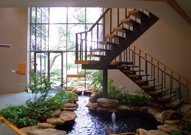 Indoor atrium with a water feature, rocks, plants, and a wooden staircase.