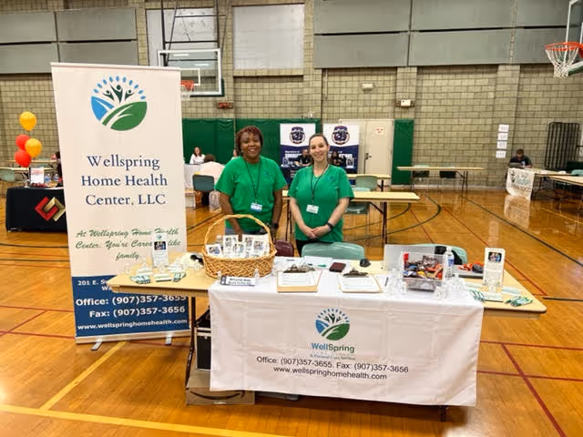 Two Wellspring representatives stand behind a Wellspring Home Health Center booth in a gym.