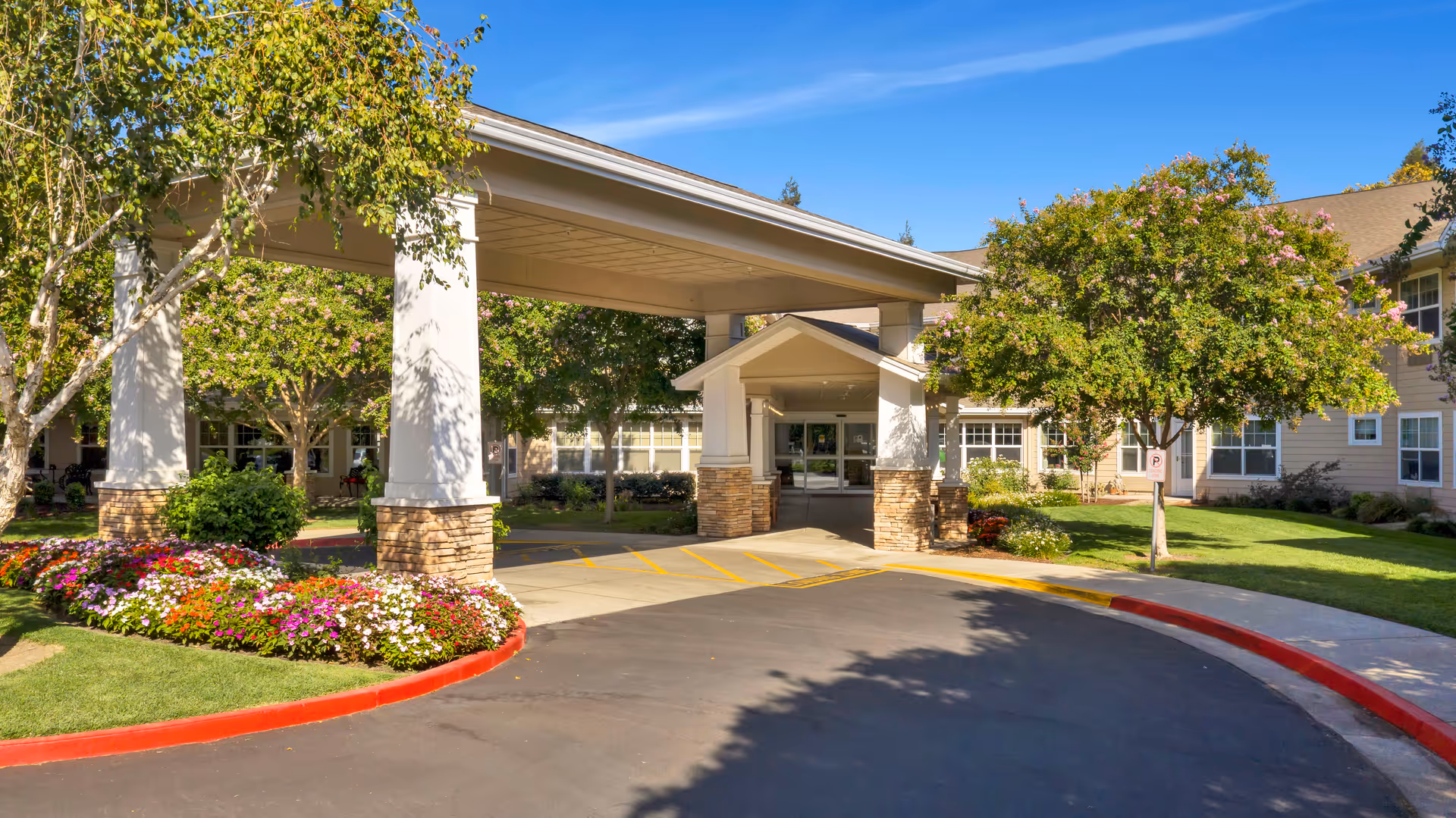 Senior living facility entrance with a covered driveway, landscaped garden, and blue sky.