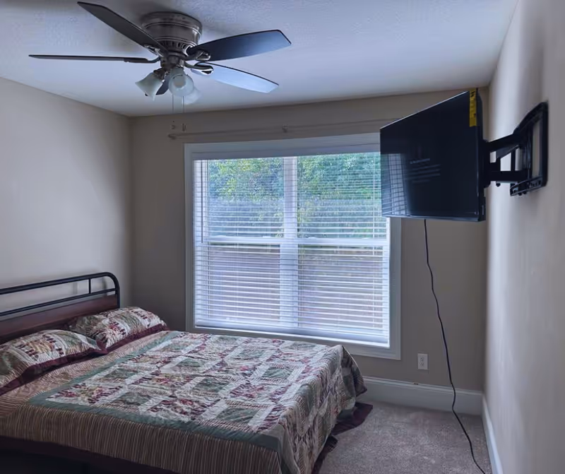 Small bedroom with a quilted bed, ceiling fan, and wall-mounted TV facing the window.