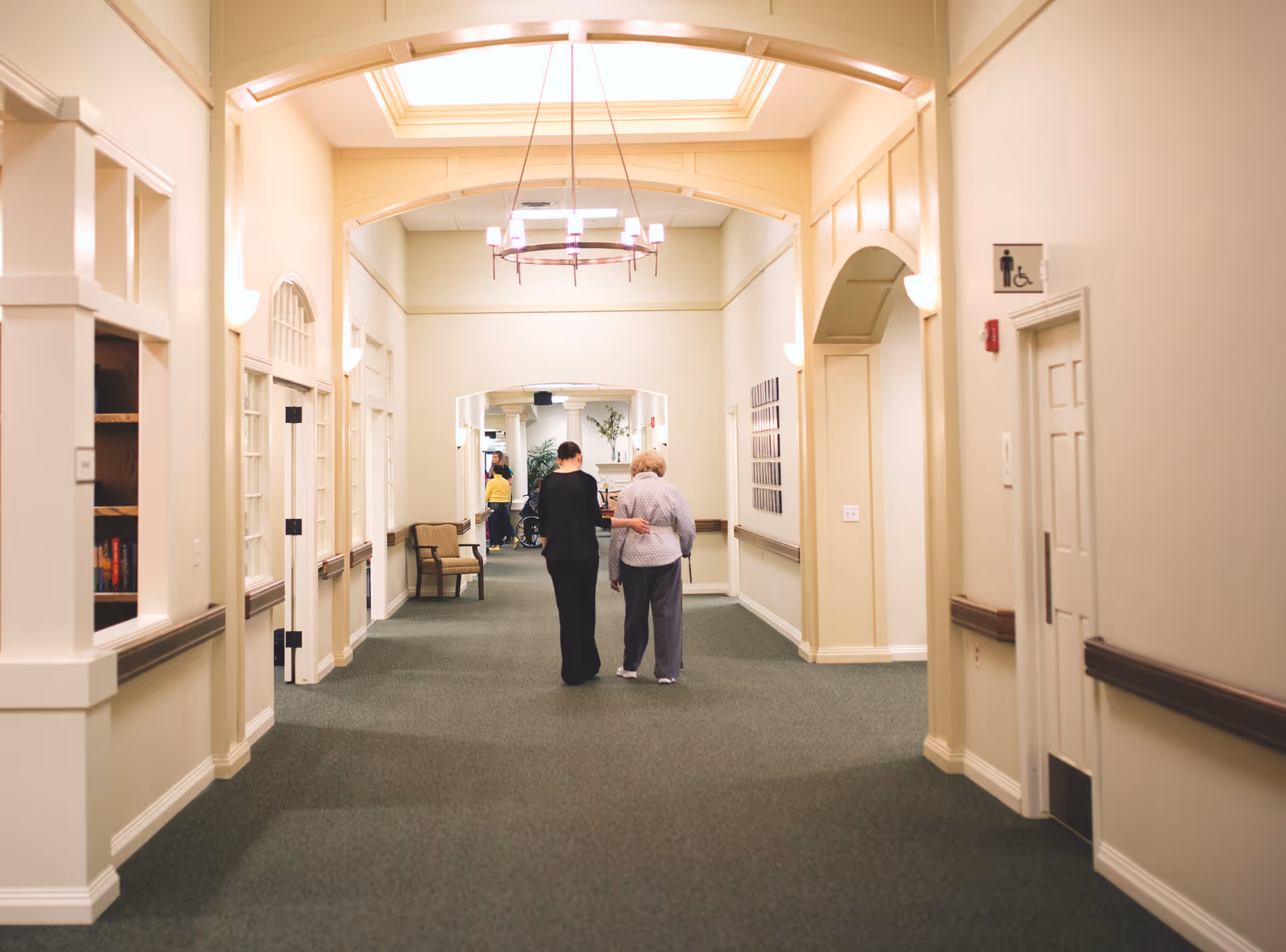 Well-lit interior hallway in a senior living facility with two people walking.