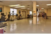 A spacious indoor fitness area with exercise machines, seating, and residents in a senior facility.