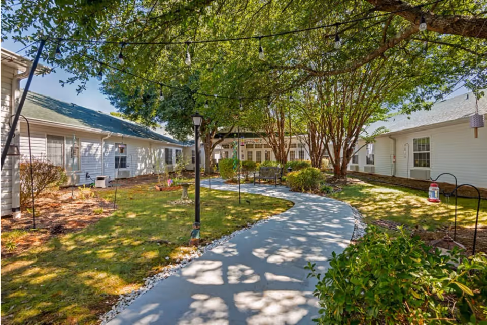 A peaceful outdoor courtyard at Brookdale Ebenezer Road featuring a winding concrete pathway surrounded by green grass, bushes, and trees providing shade. White buildings with windows and air conditioning units border the courtyard. String lights are hung above the pathway, and benches are placed along the path for seating.