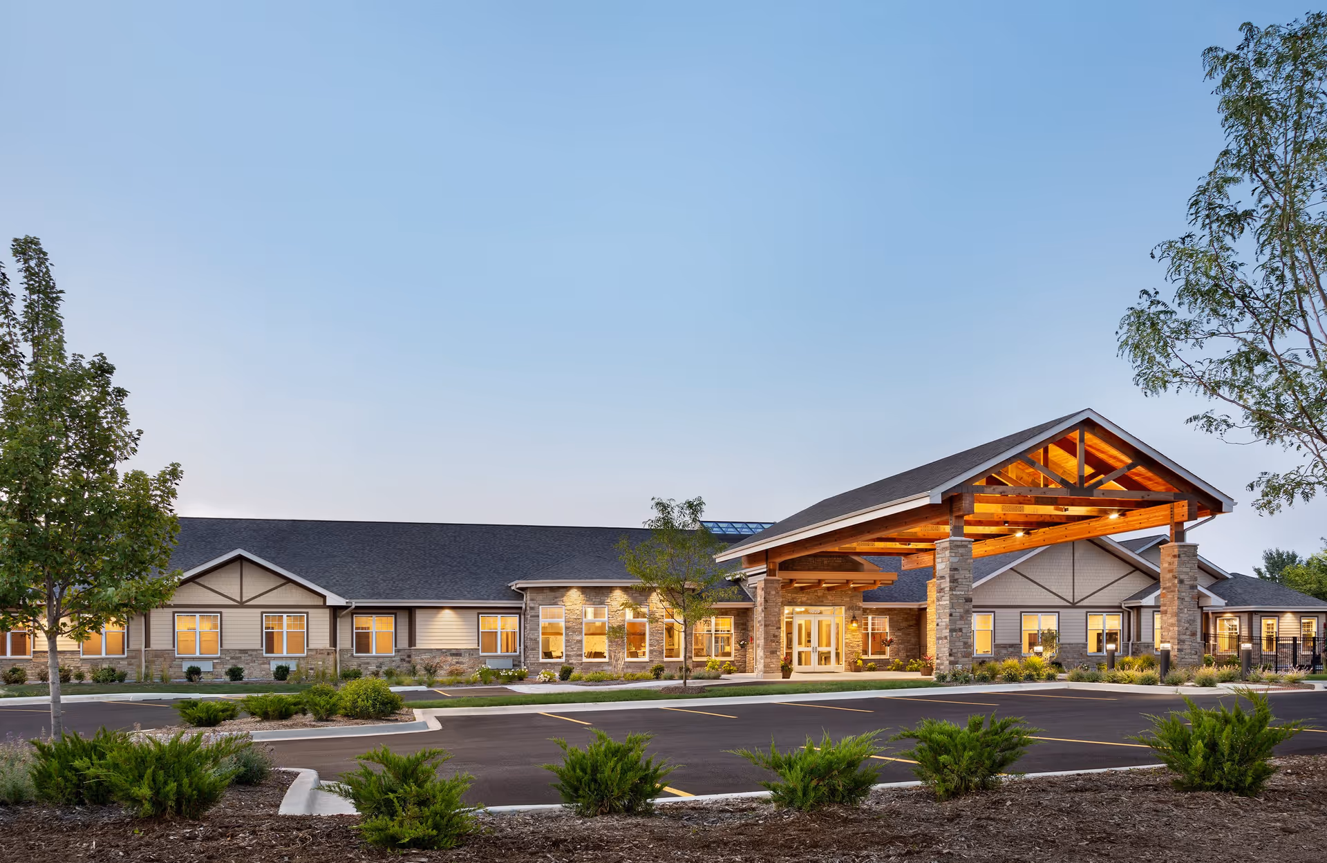 Front exterior of a single‑story senior living building with a lit wooden entrance canopy, parking area, and landscaping at dusk.