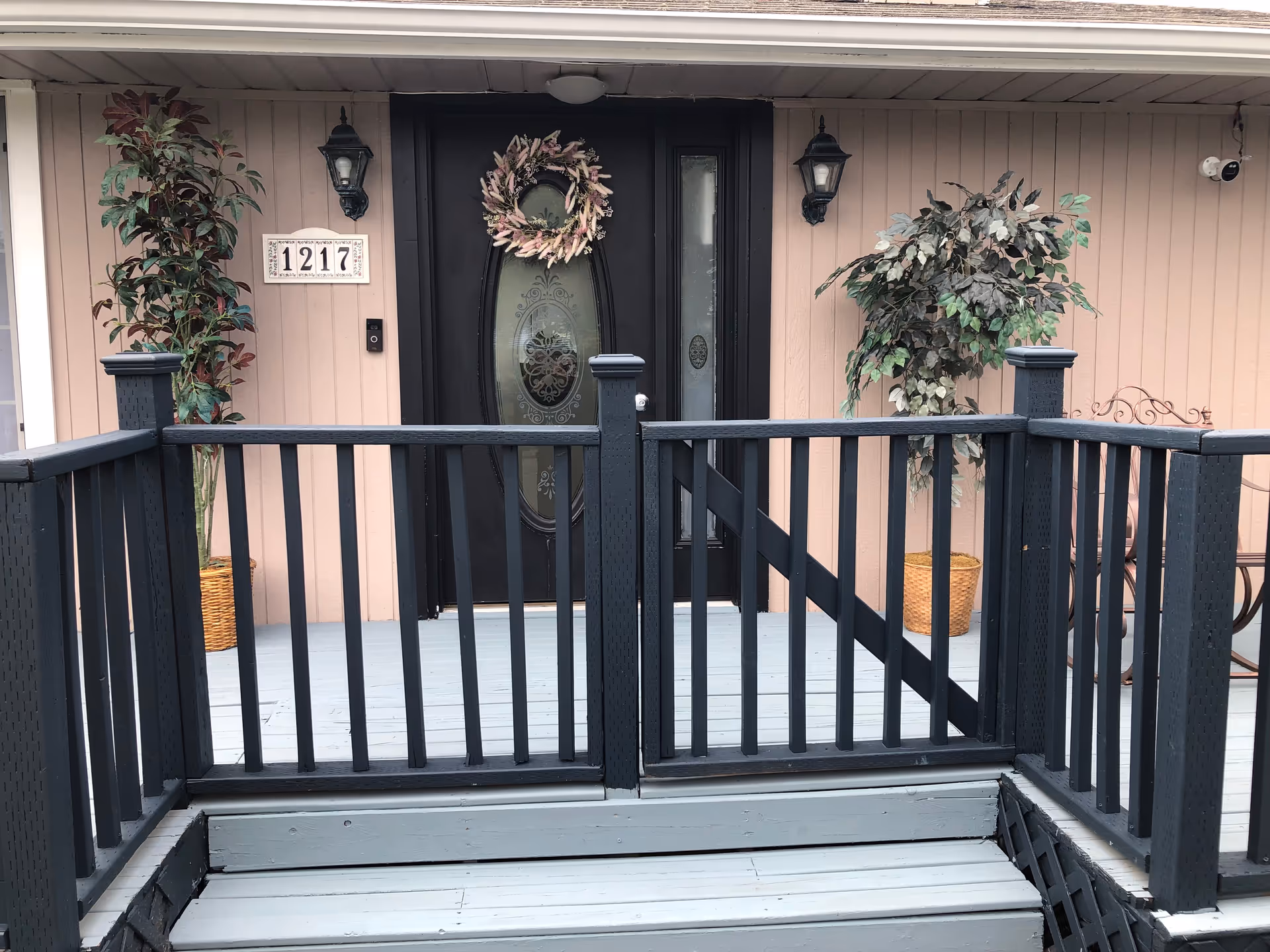 Front entrance of a residential building with wreath, lanterns, steps, and potted trees.