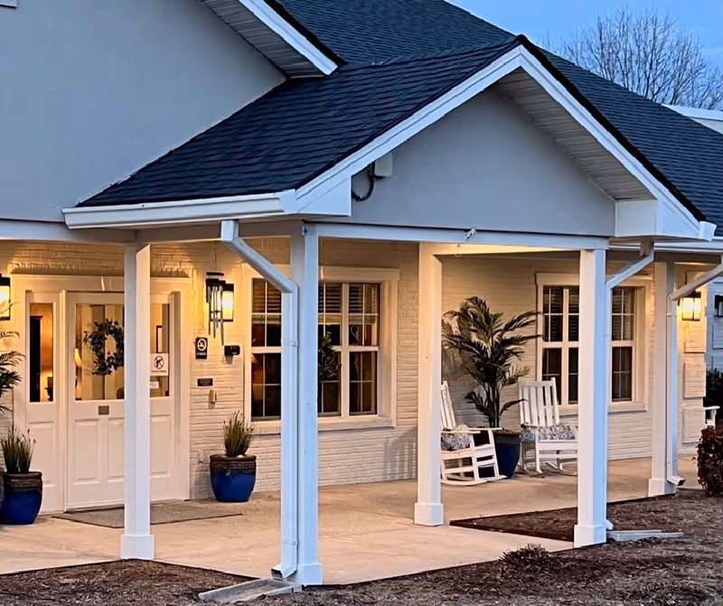 Covered front entrance of a building with white columns, rocking chairs, potted plants, and warm exterior lighting.