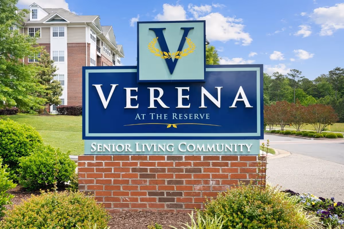 Outdoor image of a large sign for Verena at the Reserve Senior Living Community, with a multi-story residential building and greenery in the background under a partly cloudy sky.