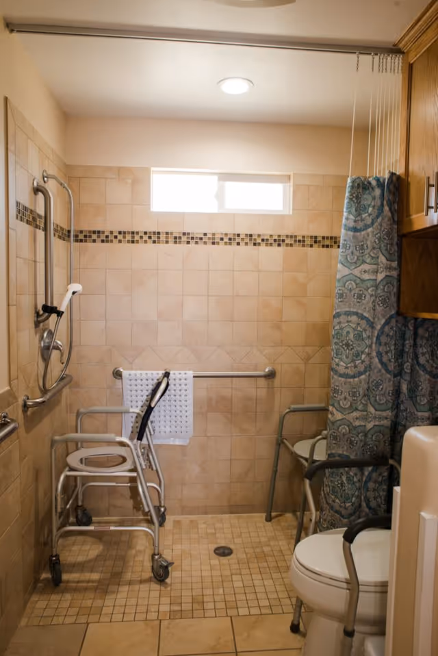 Accessible bathroom with shower chair, walker, grab bars, and a blue patterned shower curtain.