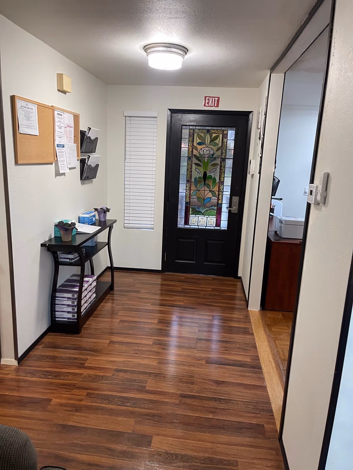 Hallway interior of a senior living facility with bulletin board, table, and stained glass exit door.