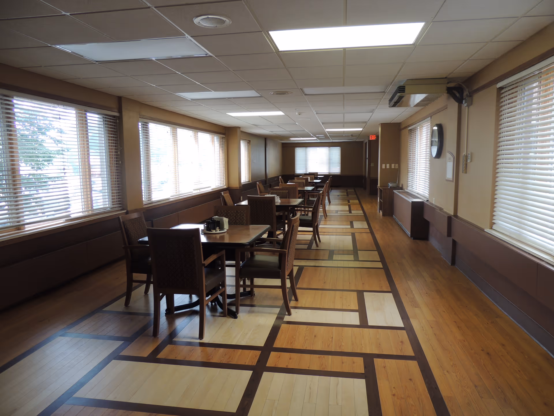 A long dining room with multiple wooden tables and chairs arranged in rows. Large windows with blinds line both sides of the room, allowing natural light to fill the space. The floor features a patterned wood design, and the ceiling has recessed lighting panels. The room appears clean and well-maintained.