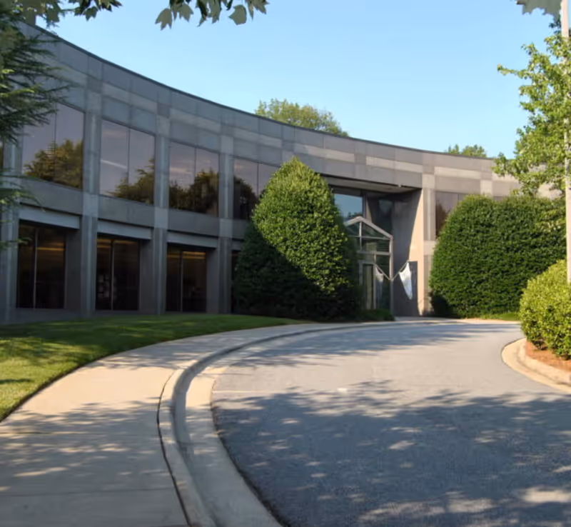 Curved modern facility entrance with glass windows, surrounded by trimmed evergreens and a paved driveway.