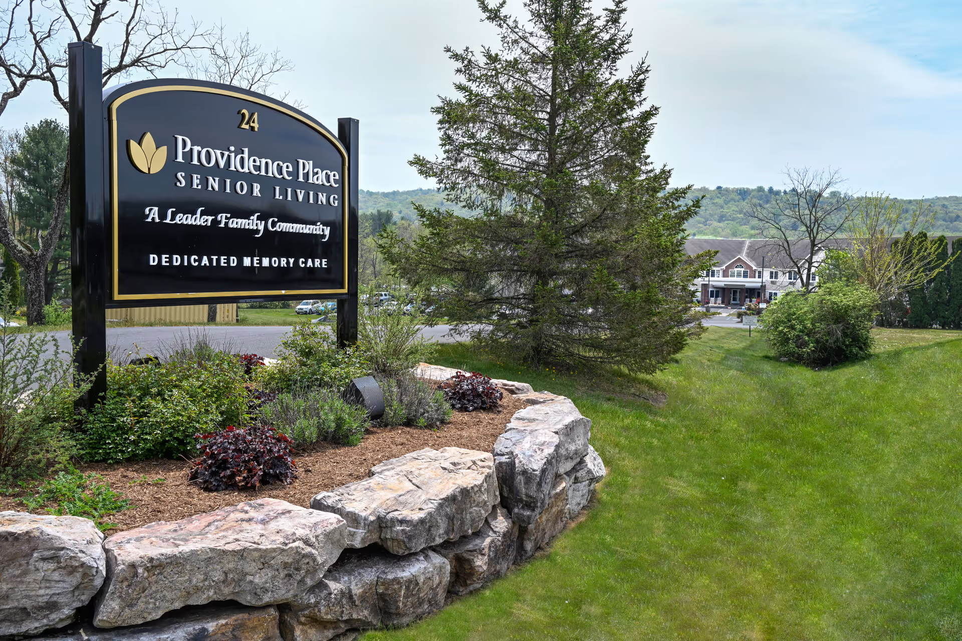 Outdoor view of Providence Place Senior Living of Pine Grove sign with landscaping including bushes, flowers, and large rocks in the foreground. A large tree and a senior living facility building are visible in the background under a cloudy sky.