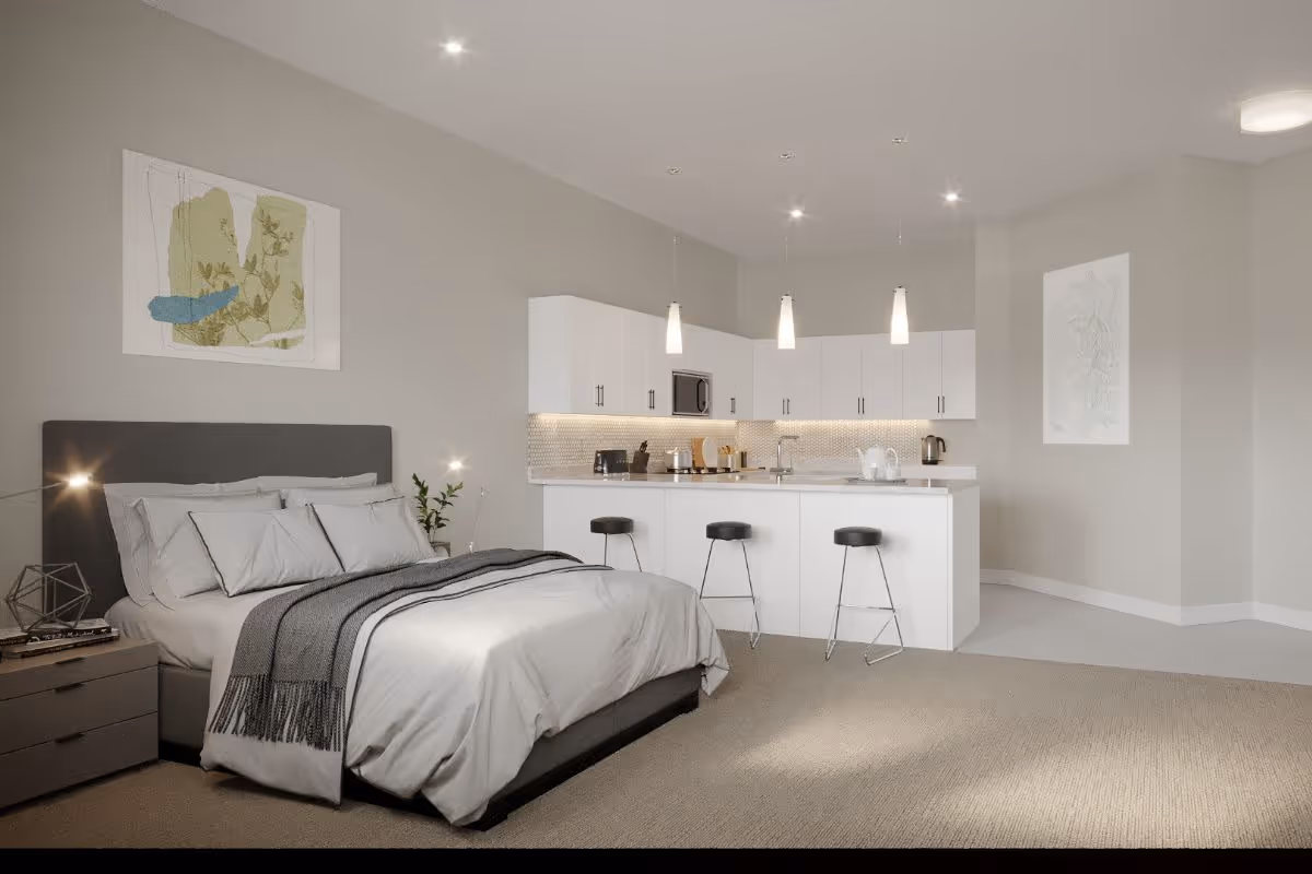Modern studio suite with a bed, kitchenette, and bar stools in soft gray tones.