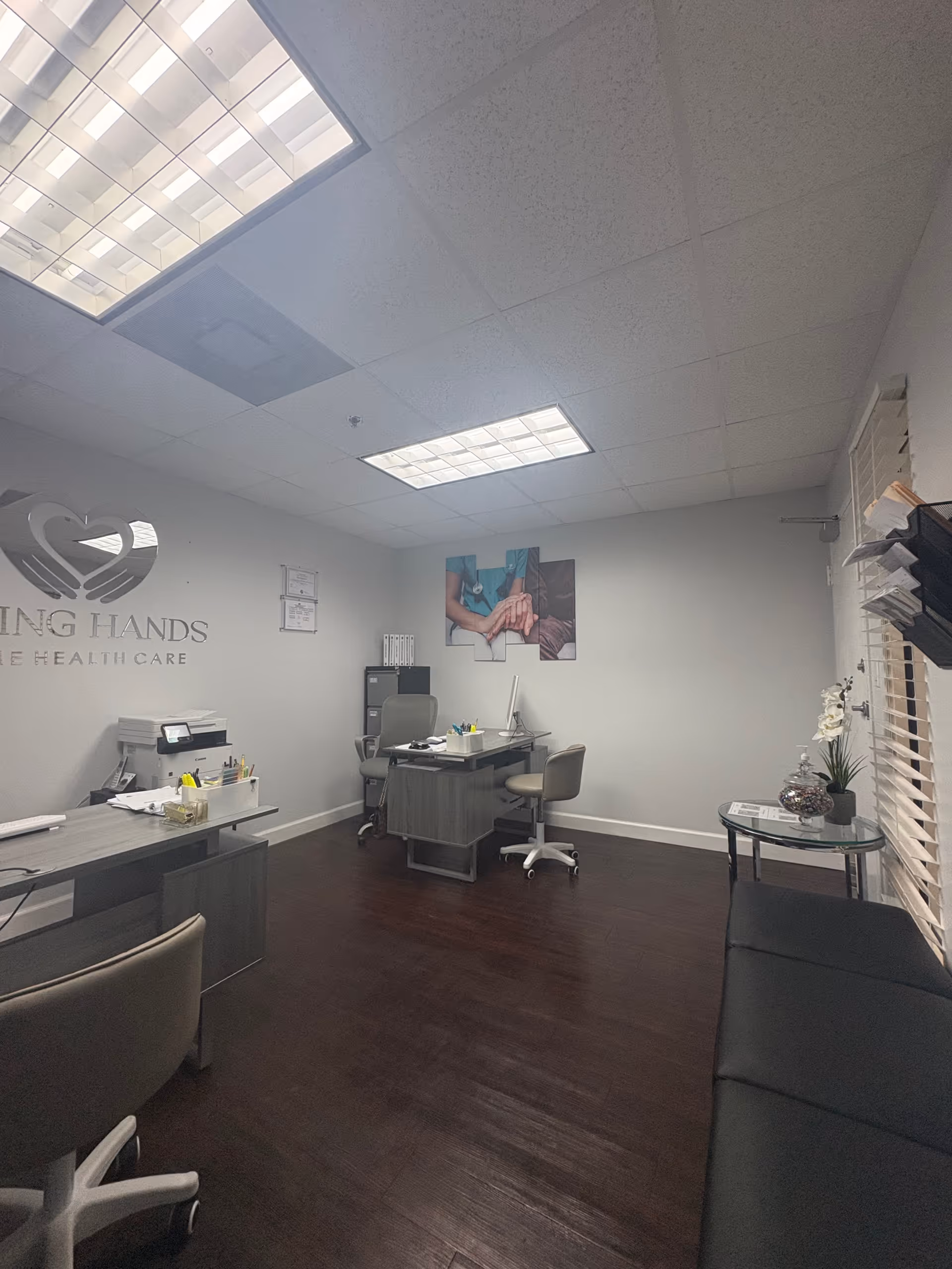Office reception area with desks, chairs, computer, and Loving Hands Health Care logo on wall.