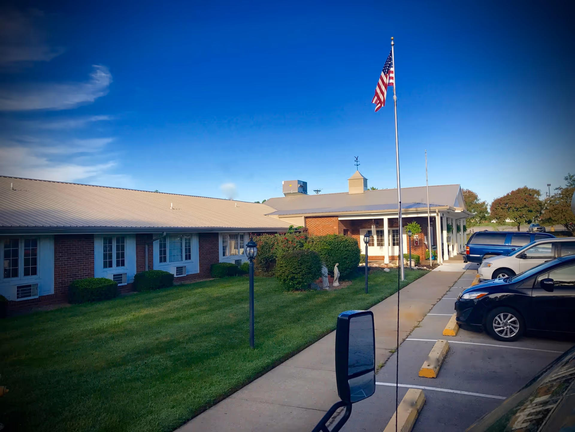 Exterior view of a single-story brick building with a metal roof, surrounded by a well-maintained lawn and bushes. An American flag is flying on a flagpole near the entrance, and several cars are parked in the adjacent parking lot under a clear blue sky.