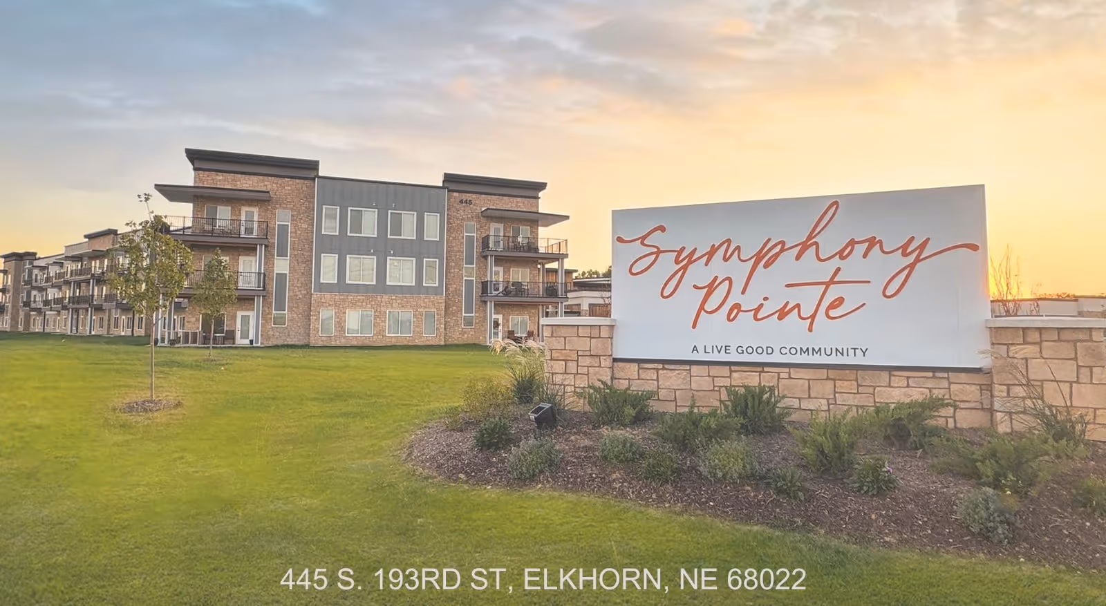 Exterior view of the Symphony Pointe sign in front of a multi-story residential building and grassy lawn at sunset.