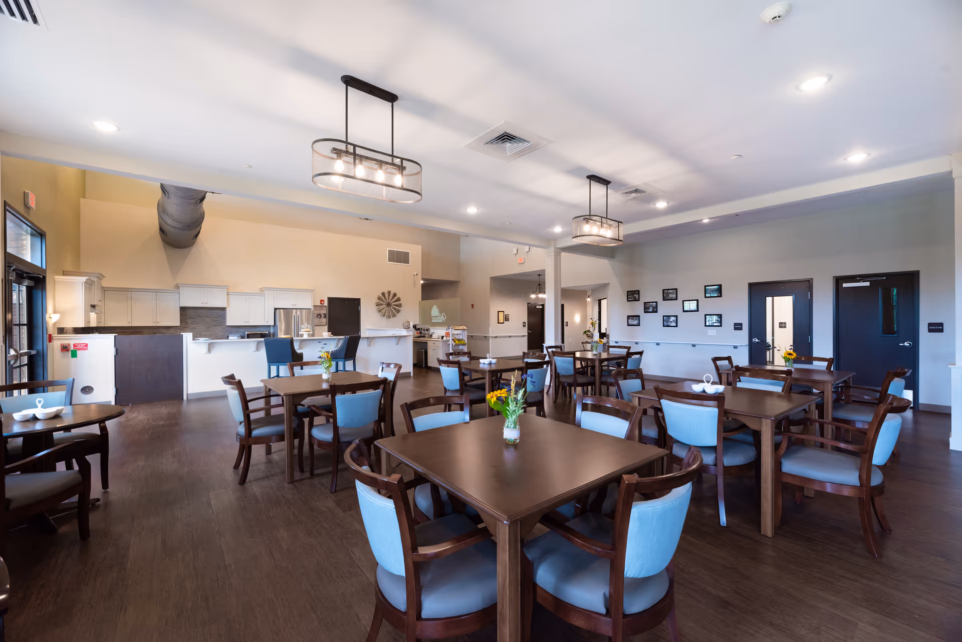A spacious dining room in an assisted living facility with multiple wooden tables and chairs featuring light blue cushions. The room has modern light fixtures hanging from the ceiling, a kitchen area with white cabinets and stainless steel appliances in the background, and framed pictures on the wall.