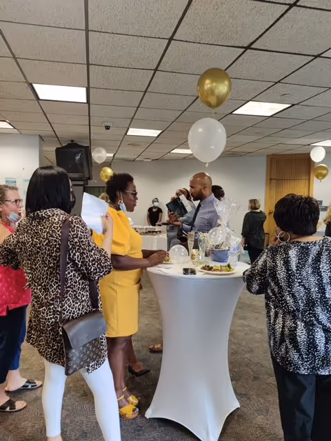 A group of people socializing around a tall cocktail table covered with a white tablecloth in a decorated indoor space. There are gold and white balloons tied to the table and ceiling. Some people are holding plates with food and drinks, and others are engaged in conversation.