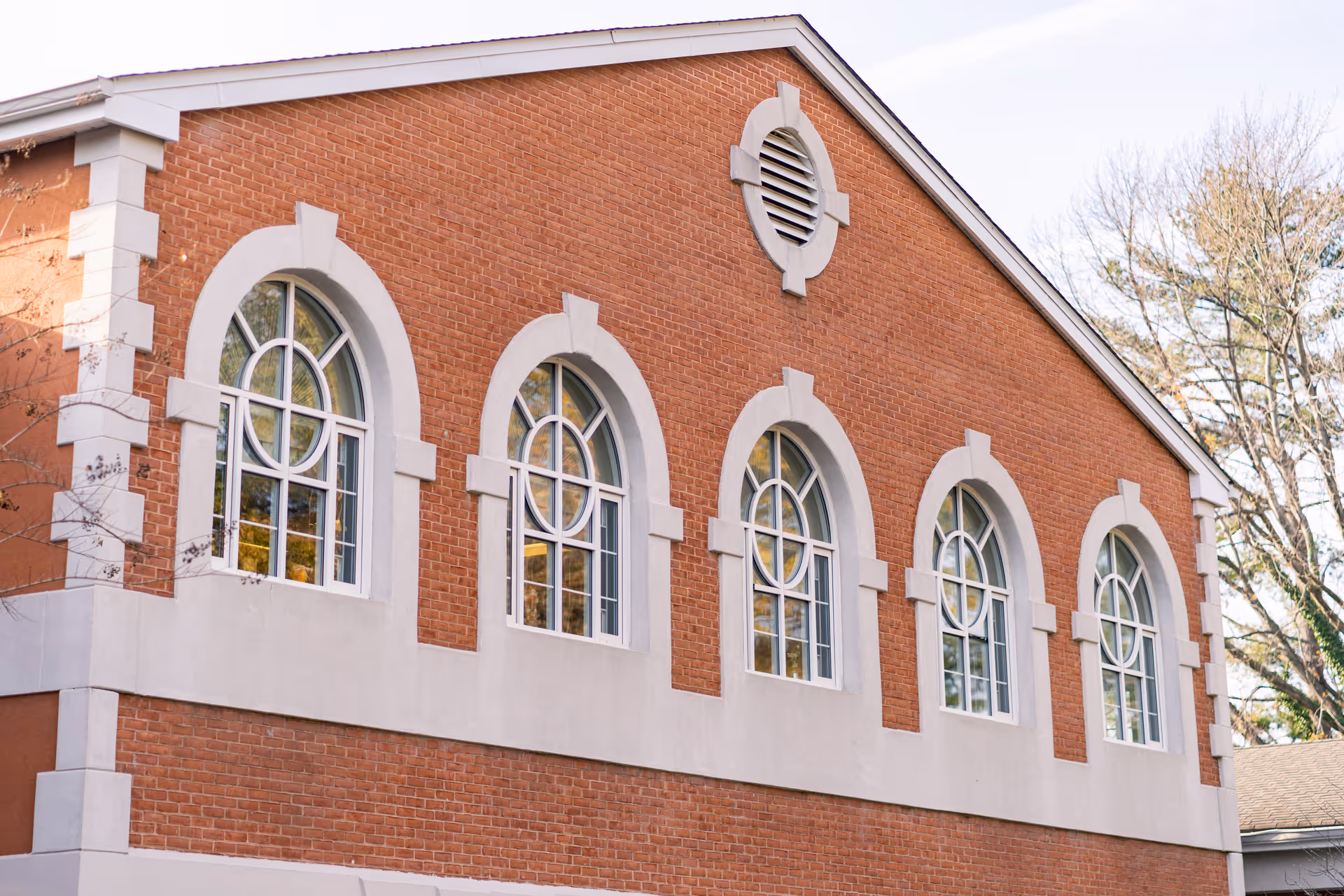 Exterior view of a brick building with five arched windows featuring decorative white trim and a small circular vent near the roof peak. Trees are visible in the background.