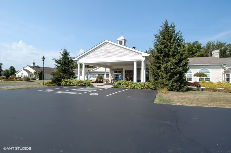 Exterior view of Whispering Pines Village showing the main entrance with a covered drop-off area supported by white columns. The building is light-colored with large windows and surrounded by greenery and trees. There is a paved parking lot with marked handicap parking spaces in front.