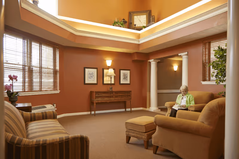 A cozy living room area with warm brown walls and carpet, featuring a striped sofa, a cushioned armchair with an ottoman, and a wooden console table against the wall. A woman with gray hair wearing a green jacket is sitting on the armchair reading a book. The room has large windows with wooden blinds, decorative columns, framed artwork, and soft lighting from wall sconces and a table lamp.