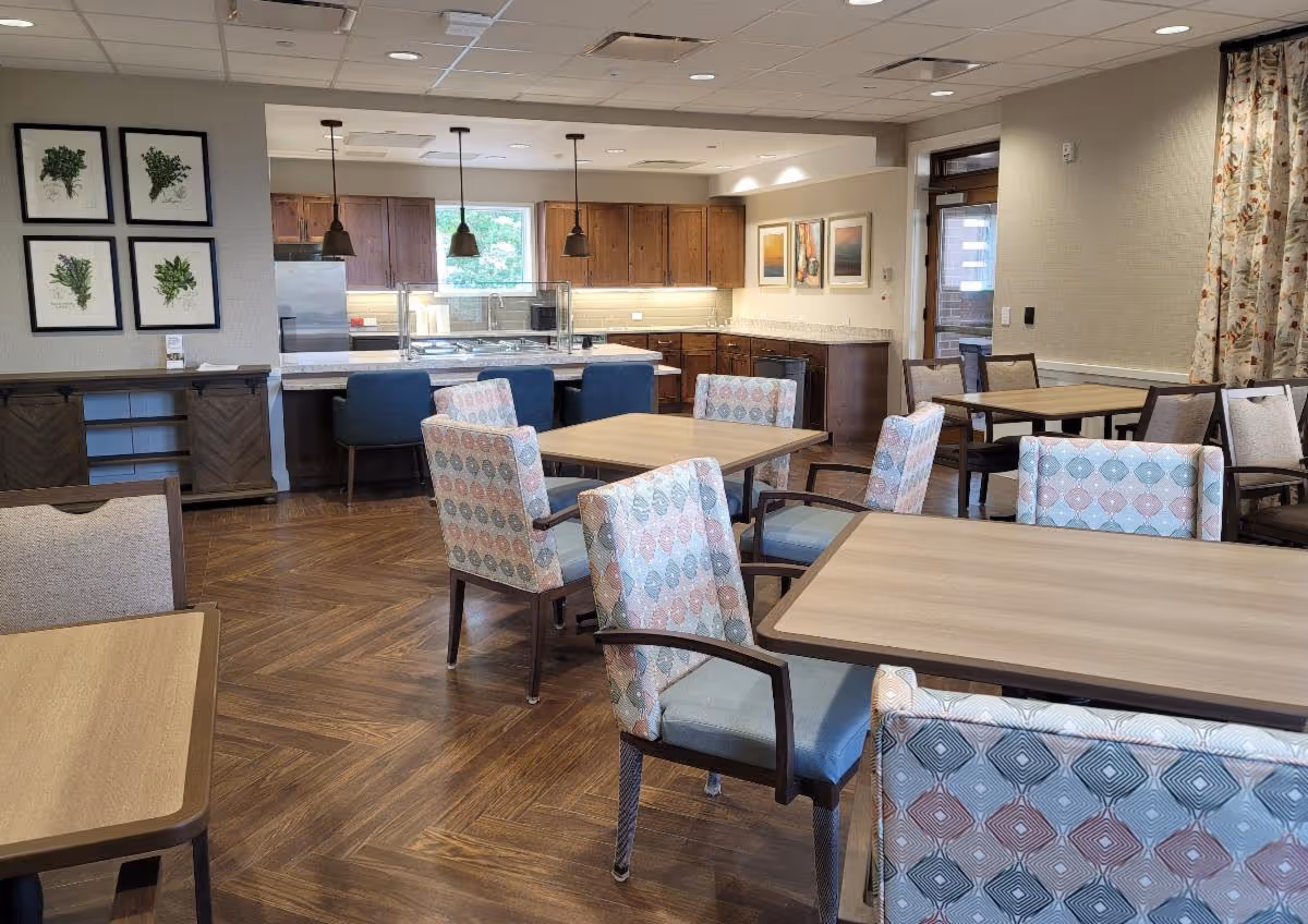 Dining room with several tables and patterned upholstered chairs facing a kitchen service counter with pendant lights.