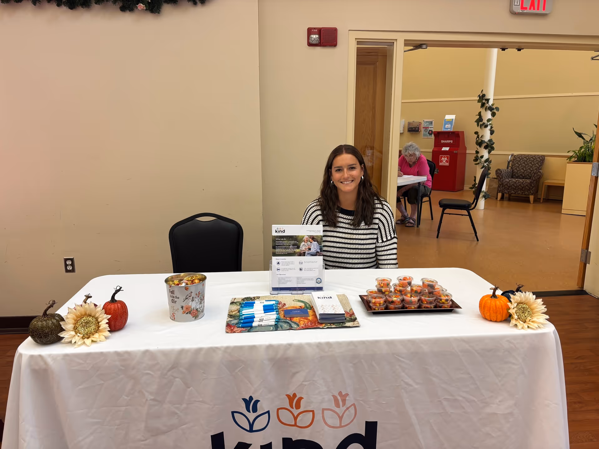 Smiling woman sits at a table with snacks and fall decor inside a senior care facility.