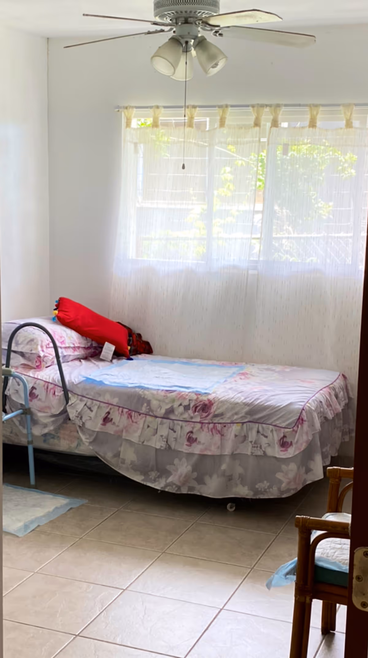 A small bedroom with floral bedspread, window curtains, a ceiling fan, and a chair.
