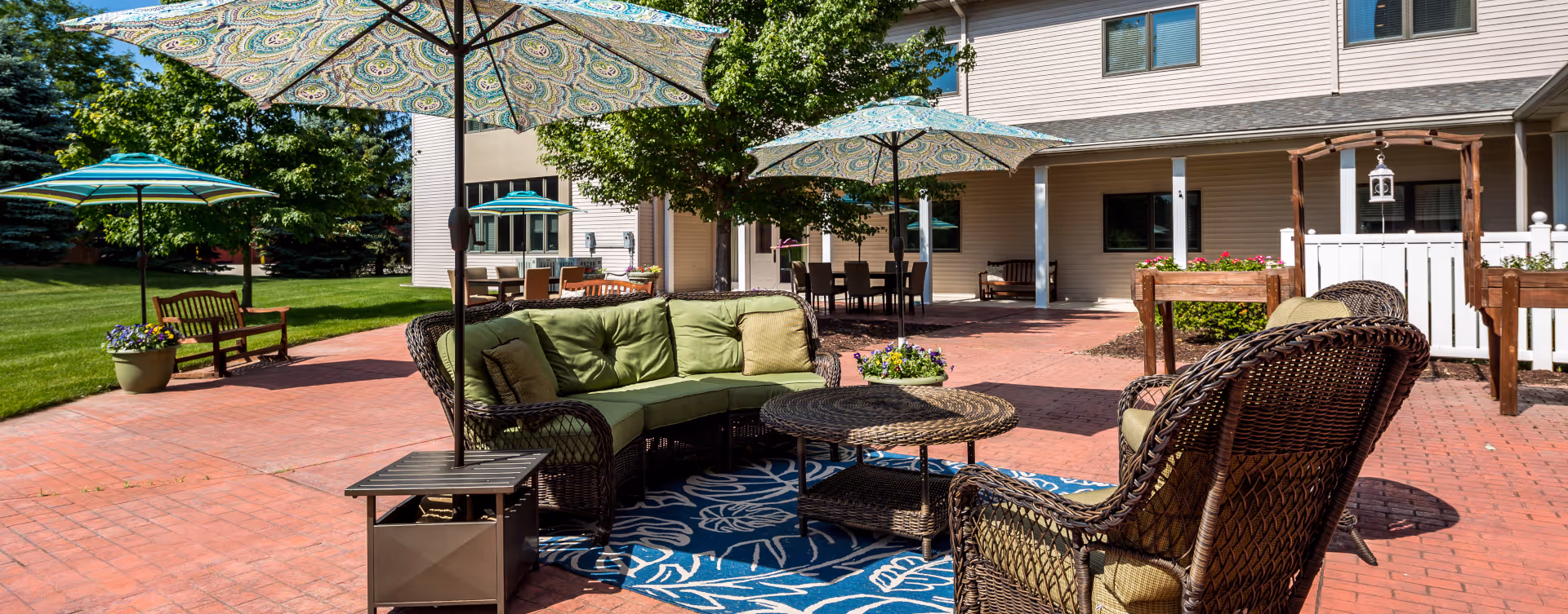 Outdoor patio area at Delta Retirement Center featuring wicker furniture with green cushions, a round wicker coffee table, patterned umbrellas providing shade, a blue and white outdoor rug, potted plants, and a building with windows and a covered porch in the background.