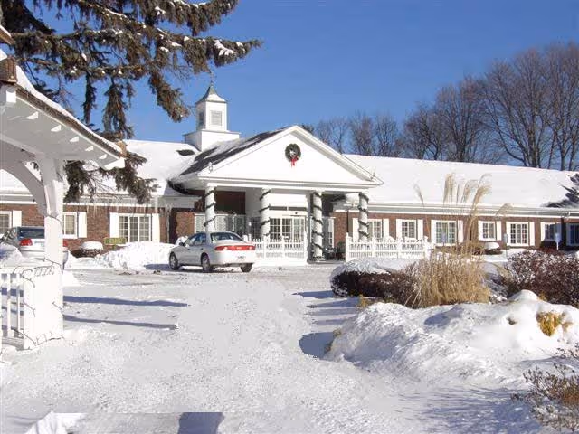 Snow-covered front entrance of a single-story brick senior living building with columns, a cupola, and a car in the driveway.