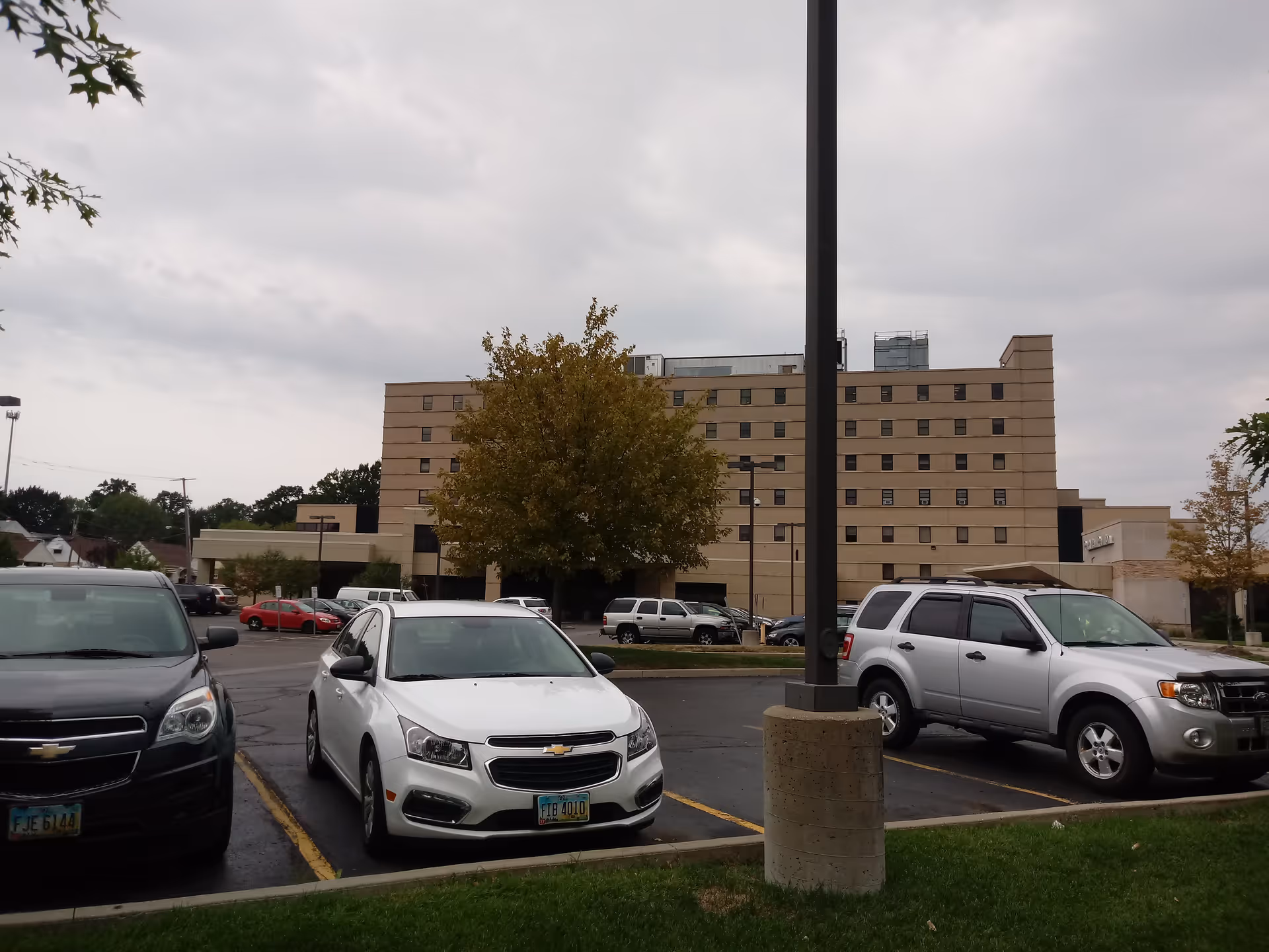 Parking lot in foreground with a large tan nursing facility under cloudy sky.