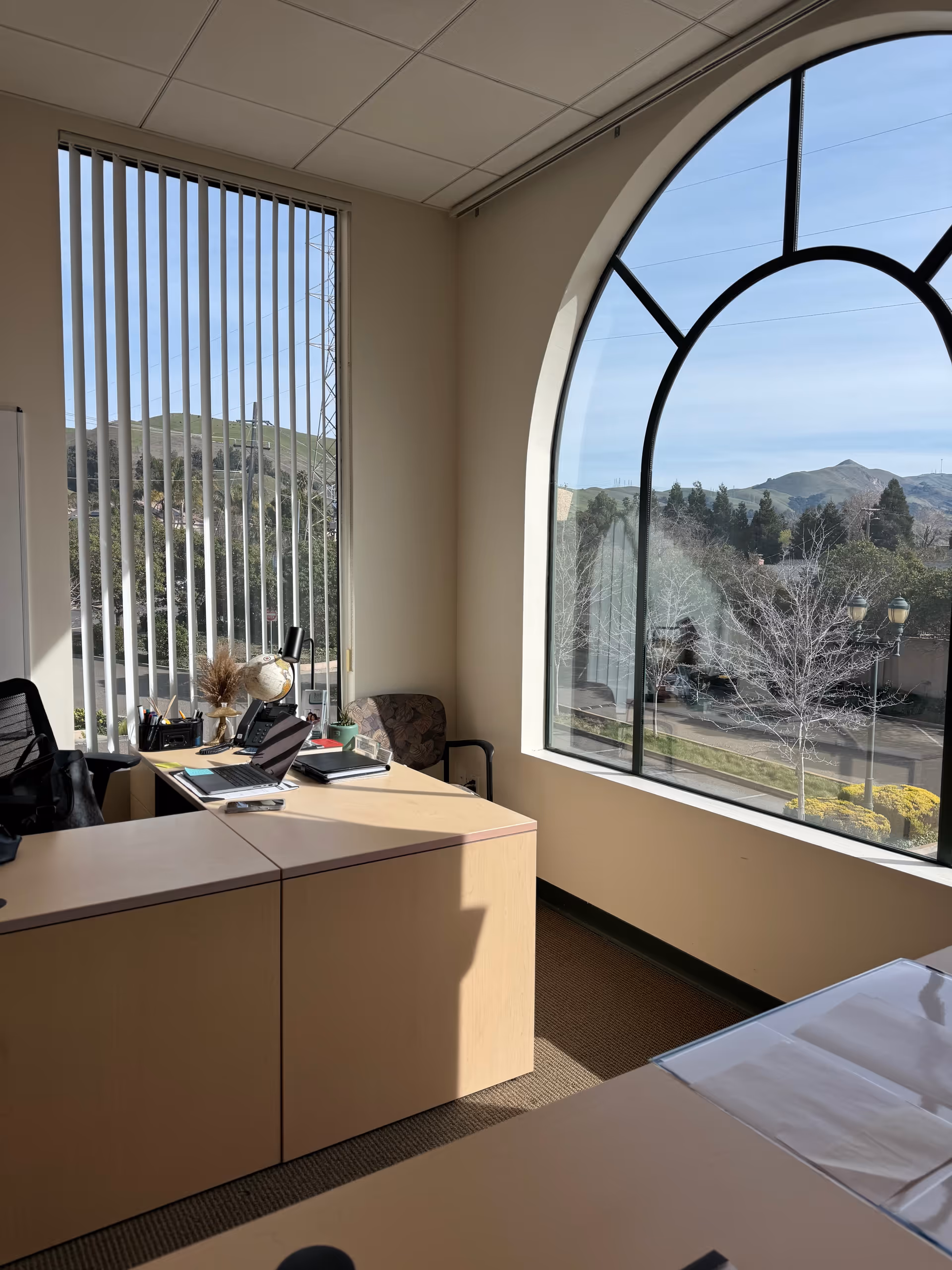 Office desk and chair beside a large arched window overlooking hills; laptops and supplies.