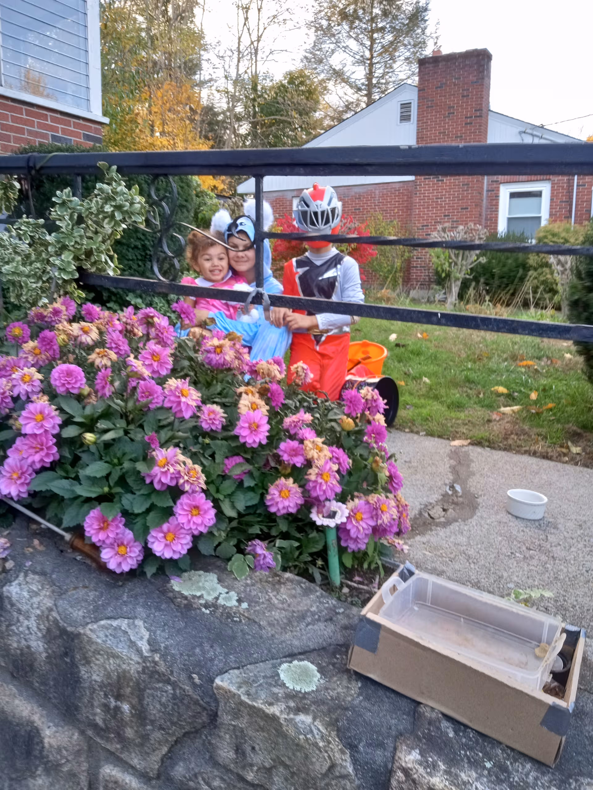 Three costumed children pose by a colorful flower bed behind a black fence outside Carlyle House.