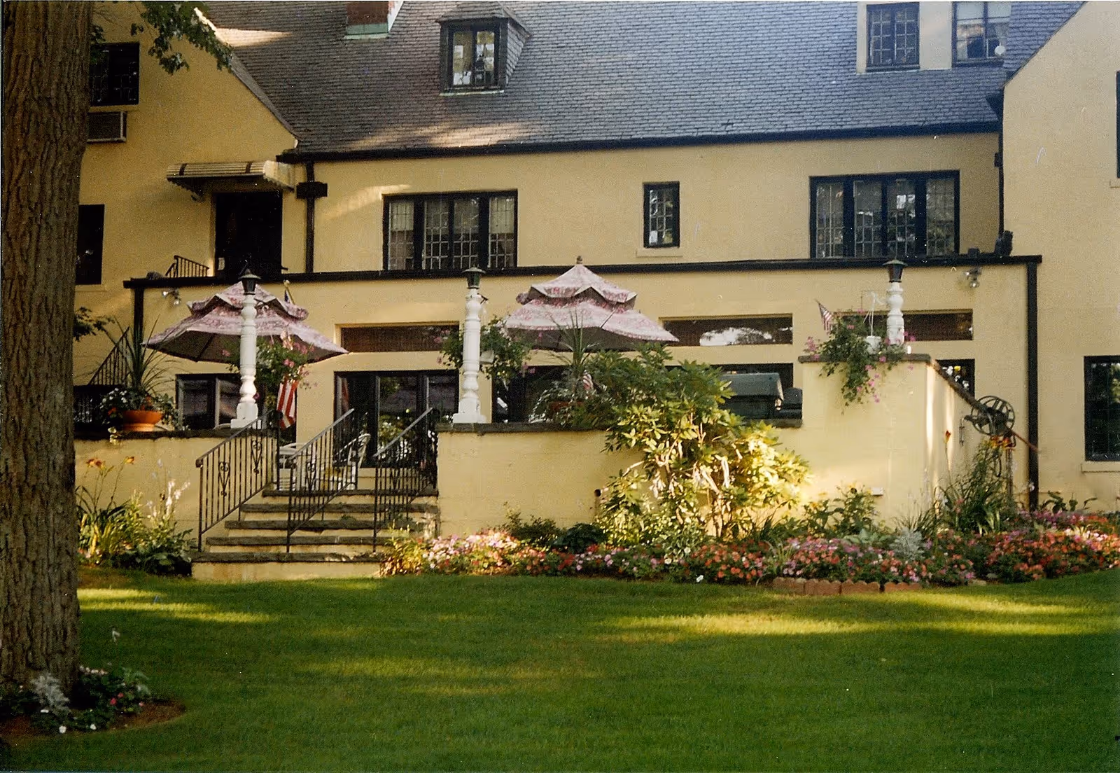 Exterior view of a large yellow building with multiple windows and a slate roof. In front of the building is a raised patio area with stairs leading down to a well-maintained lawn and garden filled with colorful flowers and shrubs. The patio has decorative white pillars and pink umbrellas providing shade.