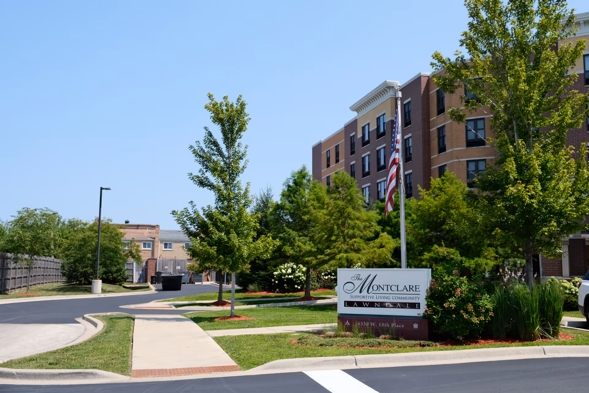 Exterior front view of The Montclare Supportive Living Community building with a sign, flagpole, trees, and a sidewalk.