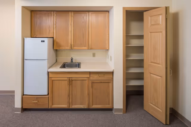 Small kitchenette with oak cabinets, a sink and mini-refrigerator beside an open pantry door.