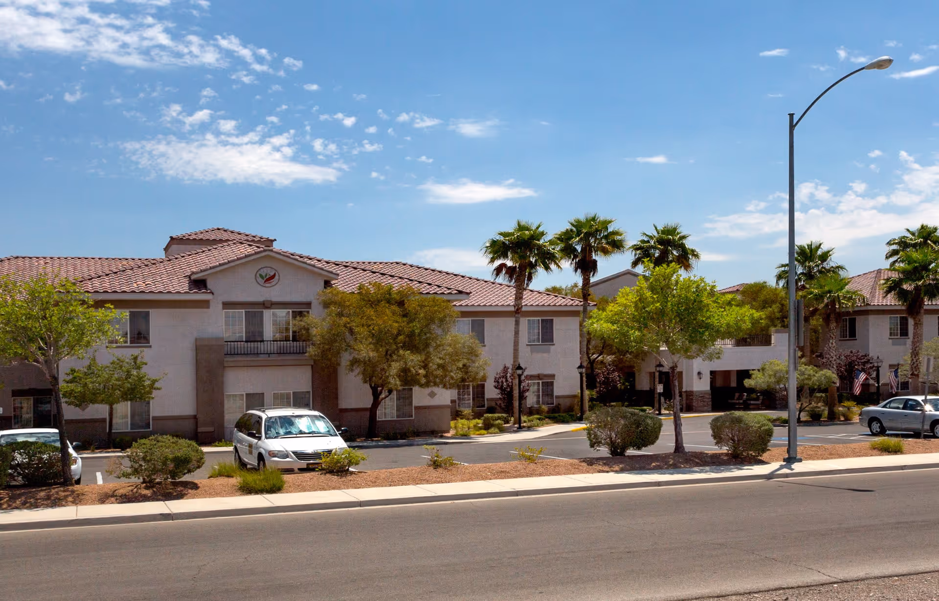 Exterior view of a two-story senior living facility building with a tiled roof, several windows, and a parking area in front. The scene includes palm trees, other greenery, and a clear blue sky with some clouds.