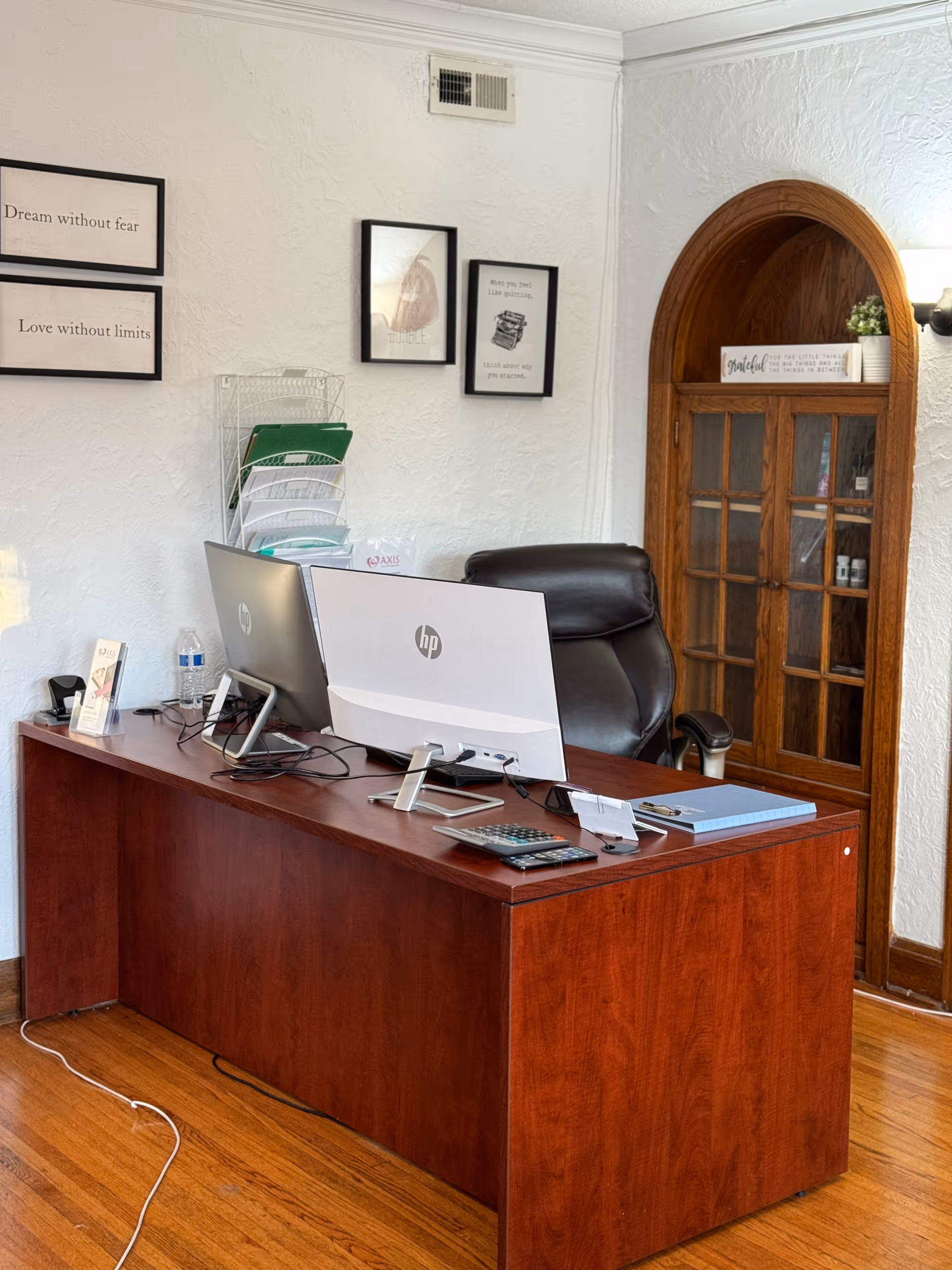 Office desk with dual computer monitors, a leather chair, and a wooden arched cabinet.