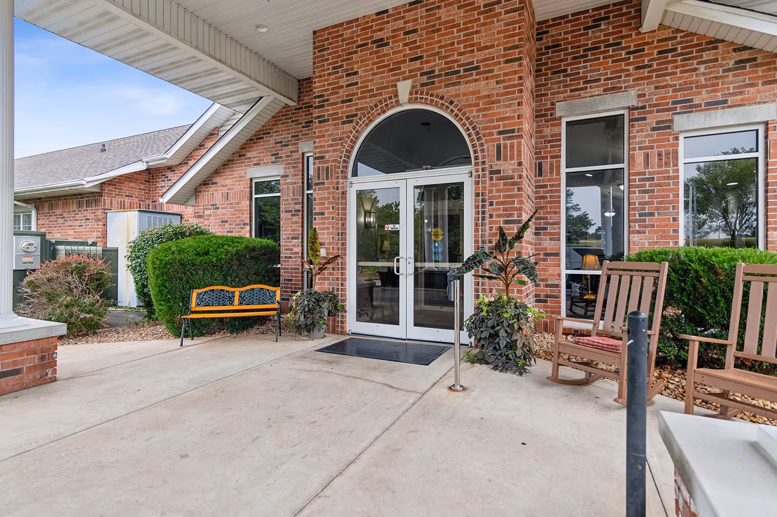 Covered brick building entrance with double glass doors, benches, plants, and rocking chairs on a concrete porch.