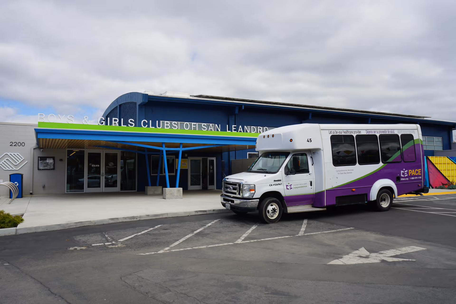 Exterior view of a blue building with glass doors and a parked white-purple bus.