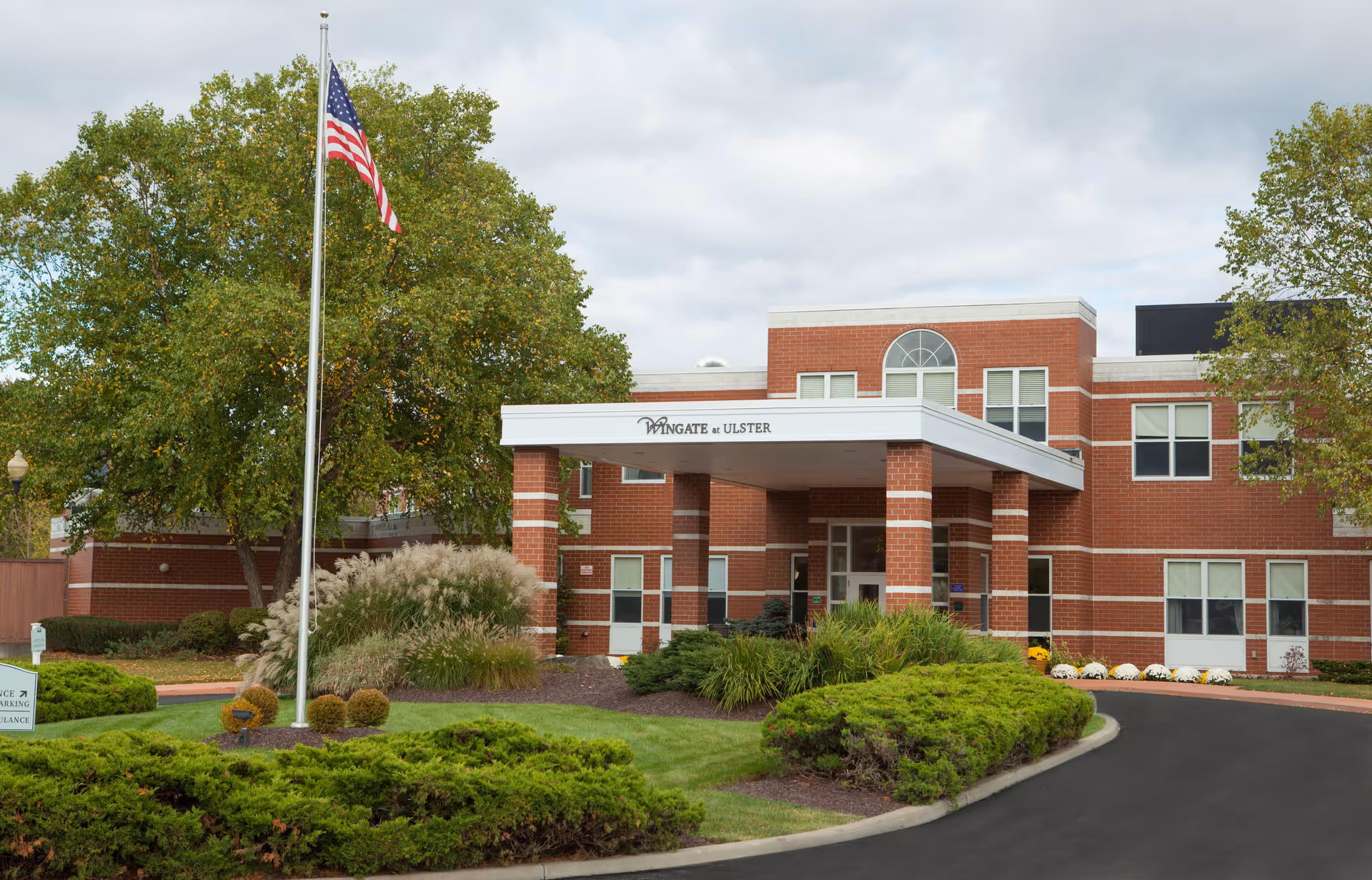 Front exterior view of Wingate at Ulster, a red brick building with white trim. There is a covered entrance supported by brick columns, an American flag on a flagpole, and well-maintained landscaping with green bushes and trees under a cloudy sky.