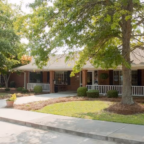 Exterior view of a single-story brick building with a covered porch, white railings, and several windows. There is a large tree with green leaves in the foreground, along with neatly trimmed bushes and a small grassy area. A concrete sidewalk runs in front of the building.