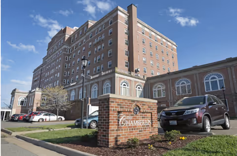 Exterior view of The Chamberlin, a large multi-story brick building with arched windows. In front of the building is a brick sign with the name 'The Chamberlin' and a parking area with several cars parked. The sky is clear and blue.