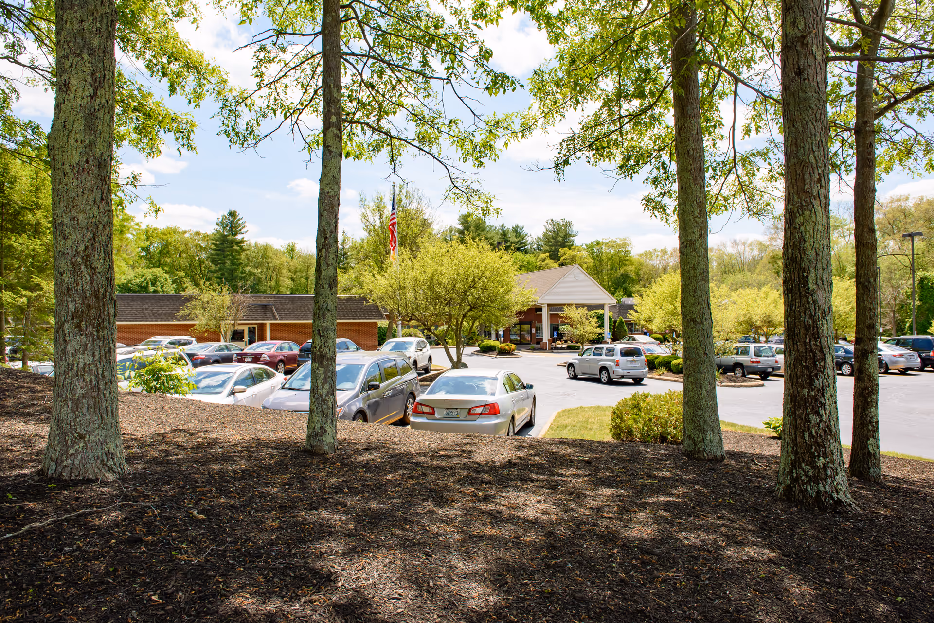 Front entrance and parking lot of a senior living facility visible through trees and landscaped grounds.