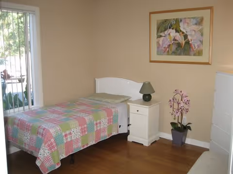 A small bedroom with a single bed covered in a colorful patchwork quilt. Next to the bed is a white nightstand with a green lamp on it. A potted orchid plant is on the floor beside the nightstand. There is a framed floral painting on the beige wall above the nightstand. A window with vertical blinds is on the left side of the room, allowing natural light to enter. The floor is wooden.